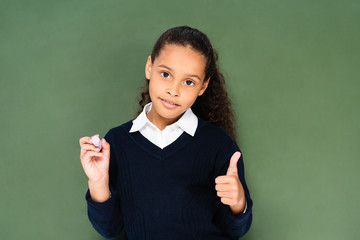 cute african american schoolgirl showing thumb up while holding near chalkboard