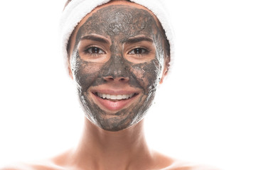 front view of smiling nude young woman in cosmetic hair band with clay mask on face isolated on white