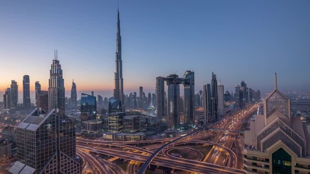 Dubai downtown skyline with tallest skyscrapers and busiest traffic on highway intersection night to day transition timelapse. Aerial view from above