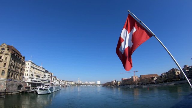 Basel, Switzerland - March 22, 2018: 4K. Swiss flag waving in the wind on the K&auml;ppelijoch bridge, which crosses the Rhein river. Buildings around the river, people walking. Switzerland.