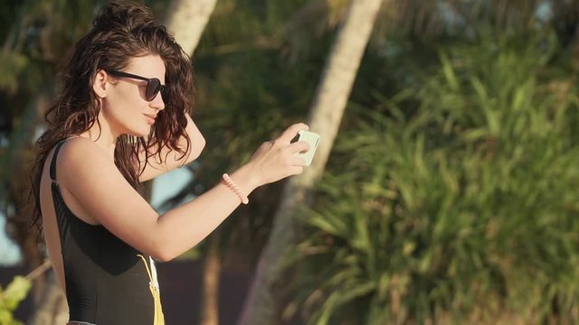Pretty young woman in sunglasses and swimsuit takes selfie on beautiful sandy beach by sea. Happy smiling girl pulls her hair, spinning and taking pictures of herself with smartphone. Slow motion.