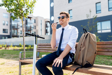 business and people and concept - young smiling businessman with backpack and scooter sitting on street bench and using voice command recorder on smartphone in city