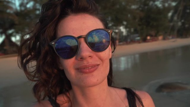 Young happy caucasian woman is spinning and dancing on beach of tropical island at sunset. Smiling pretty girl in sunglasses enjoys freedom and vacation on sandy coast by sea. Selfie POV slow motion.