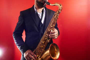 Obraz na płótnie Canvas Portrait of professional musician saxophonist man in suit plays jazz music on saxophone, red background in a photo studio