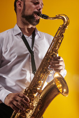 Naklejka na ściany i meble Portrait of professional musician saxophonist man in white shirt plays jazz music on saxophone, yellow background in a photo studio, side view