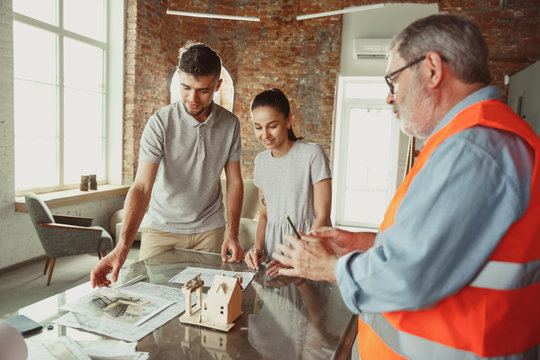 Foreman or achitect engineer shows future house, office or store design plans and model to a young couple. Meeting at the construction office to talk about facade, interior decoration, home layout.