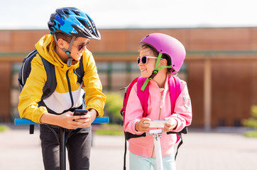 education, childhood and technology concept - happy school children in helmets with smartphones and scooters outdoors