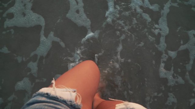 Top view close-up of young woman legs standing on sea shore at sunset. Foamy sea waves gentrly wash sand and feet, warm soft evening sun reflects on wet surface. First person view slow motion footage.