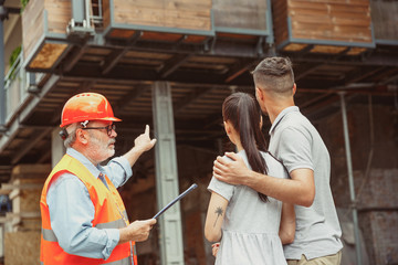 Foreman or achitect engineer shows future house, office or store design plans to a young couple. Meeting at the construction site to talk about facade appearance, interior decoration, home layout.