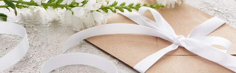 panoramic shot of beige envelope with white ribbon near flowers on textured surface