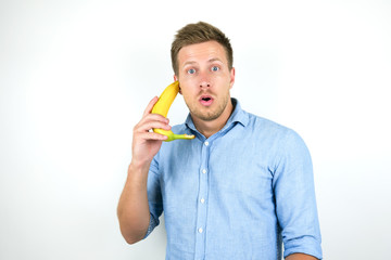 young handsome man holding one fresh banana as a phone near his ear looks surprised on isolated white background © studioprodakshn