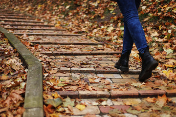 A path of yellow brick strewn with autumn leaves leads into the distance. Legs of a girl in blue jeans and boots are walking.