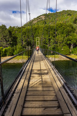 Fototapeta na wymiar Rear view of a young woman practicing hiking wearing a backpack crossing a wooden suspension bridge over a wide river in Los Alerces National Park, Patagonia, Argentina