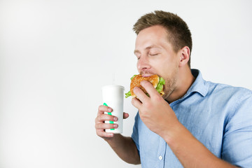 young handsome man holding soda drink and biting burger from fast food restaurant looks hungry on isolated white background