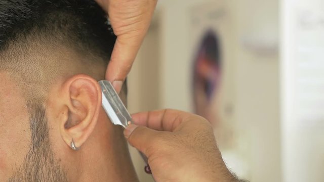 Stylish barber shaves his client neck in an old-fashion manner. Young man getting a shave with blade in traditional barbershop. Hairstylist trims customer beard with a straight razor. Close-up view