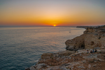 Friends seated on cliff and watching Sunset at Atlantic ocean coast, in Carvoeiro beach clifs, Algarve, Portugal.