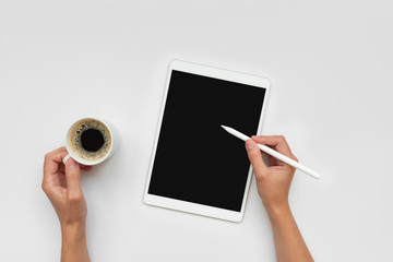 Cup of coffee and feminine hands drawing on tablet by pencil on white desk. Top view. Copy space. Mock-up