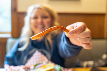 Hungry blonde woman holds up a single sweet potato french fry while eating in a restaurant. Woman...