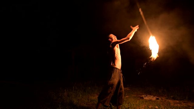 Low key. Young male with long hair and bare torso rotates burning torch outdoors on a black night video slow motion. Modern fakir does tricks with a burning staff