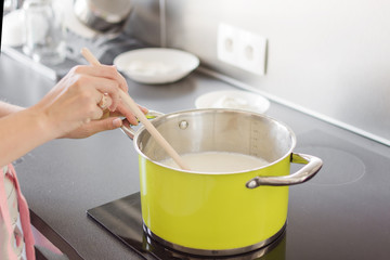 Woman preparing bechamel sauce or cream in a pan. Woman hand is mixing boiling milk with wooden...