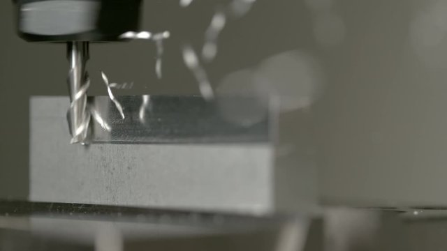 SLOW MOTION, MACRO, DOF: Twisted pieces of excess metal fly off a piece of aluminium during lathing by a CNC drill. Fine cut end mill shaving the side of a matt block of metal. Micro machining process