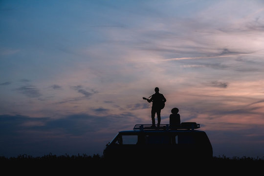 Silhouette of a man with a guitar and a woman in a hat stand on the roof of a car on the background of the sunset. Silhouette romance concept