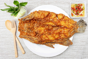 Deep fried sea bass with fish sauce and green mango chili fish sauce, based on chili fish sauce, addition of shredded green mango and lime juice serving on gray table mat with wooden spoon and fork.