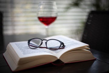 a pair of glasses lies on an open book and in the background is a wine glass