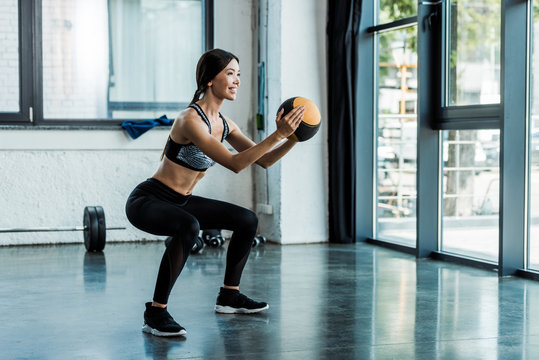 cheerful sportswoman holding ball and doing squat exercise