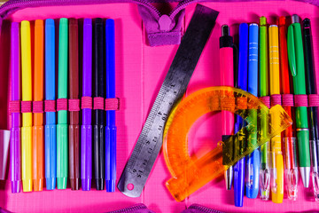 Different school stationeries (pens, pencils, felt tip pens, ruler and protractor) in a pink pencil box. Top view