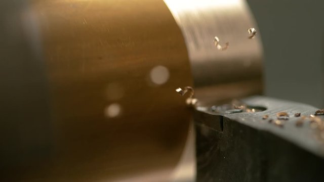 SLOW MOTION, MACRO, DOF: Automated lathing equipment carving the outer layer of a shiny brass workpiece. Sharp CNC machine blade cuts into a golden cylinder and creates twisted chips of excess metal.