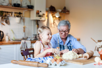Senior woman is teaching child to cook homemade bread in cozy kitchen. Grandmother and little girl are laughing together. Cute kid is helping to prepare dough. Lifestyle authentic moments.