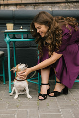 Dog and girl in street cafe. Beautiful woman in purple dress pettin small cute dog sitting under the table. Street cafe