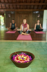 Close up of flower offering for prayer in traditional Indonesian temple in Bali with three diverse people meditating in background