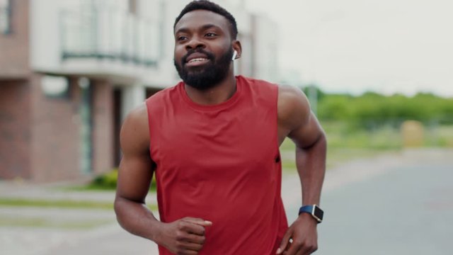 Portrait of positive afro-american running athlete man running and smiling outdoor. Attractive fit young man enjoying music jogging in modern neighbourhood on sunny day.