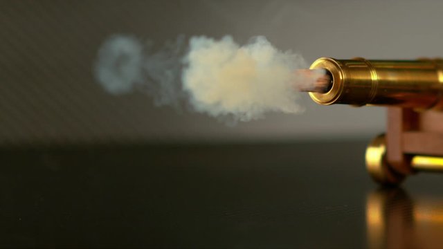 SLOW MOTION, MACRO, DOF: Shiny Vintage Cannon Emits White Smoke Before Firing A Round Shot. Retro Toy Cannon Shoots Small Bullet Across The Room. Particles Fly Out Of Barrel After Firing A Cannonball