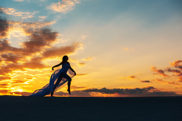 A girl in a fly white dress dances and poses in the sand desert at sunset