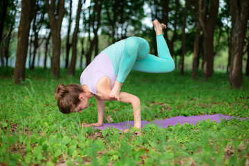 Thin brunette girl plays sports and performs beautiful and sophisticated yoga poses in a summer park. Green lush forest on the background. Woman doing exercises on a yoga mat