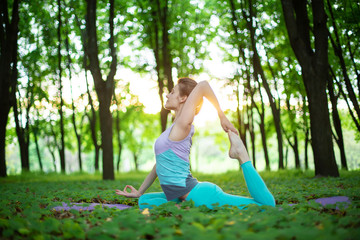 Thin brunette girl plays sports and performs yoga poses in a summer park on a sunset background....