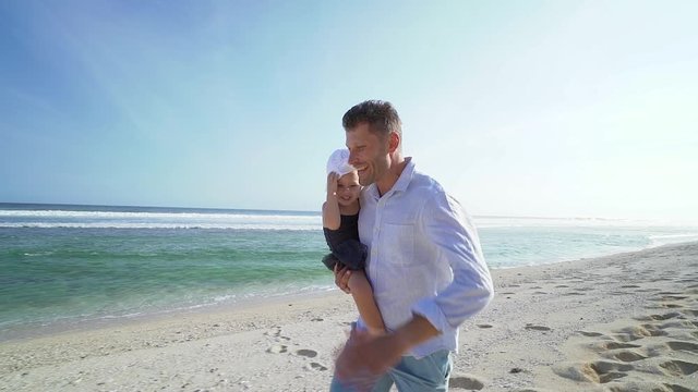 Happy smiling father holds cute daughter in hands and running on the beach at sunny day