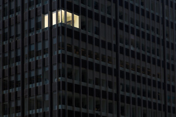 Corner office with lights on and windows glowing in a darkened office building at night indicating...