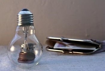 Pile of metal coins in a glass light bulb. In the background, an open wallet with paper money. Concept of saving electricity and associated finance.