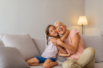 Grandmother and granddaughter hug. Little cute girl and her grandmother are spending time together at home. Having fun, hugging and smiling while sitting on sofa. - obrazy, fototapety, plakaty