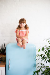 Little girl is sitting on the fridge. Sits on a beautiful retro refrigerator of blue color, stylish interior.