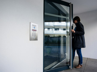 Side view of woman entering modern apartment building with glass door and modern intercom system