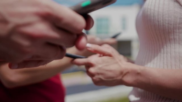 Young people standing in row showing modern mobile phones outdoors at sunlight. Happy friends busy typing on smartphone gadgets in leisure time on urban background.