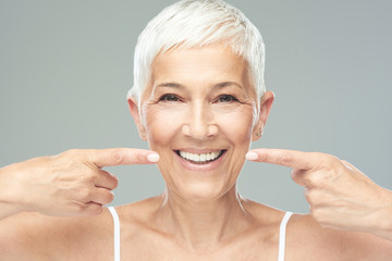 Beautiful Caucasian  smiling senior woman with short grey hair pointing at her teeth and looking at camera. Beauty photography.
