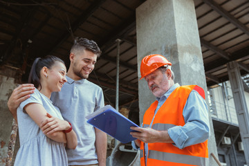 Fototapeta premium Foreman or achitect engineer shows future house, office or store design plans to a young couple. Meeting at the construction site to talk about facade appearance, interior decoration, home layout.