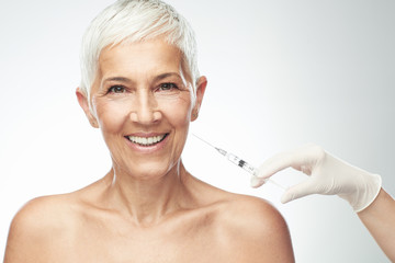 Beautiful smiling Caucasian senior woman standing in front of gray background and taking a shot of anti age product. Beauty photography.