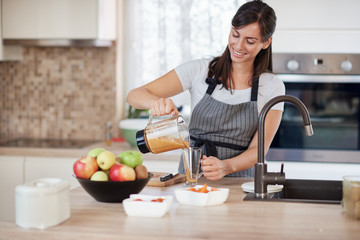 Beautiful smiling Caucasian woman in apron standing in kitchen and pouring fresh smoothie into glass.